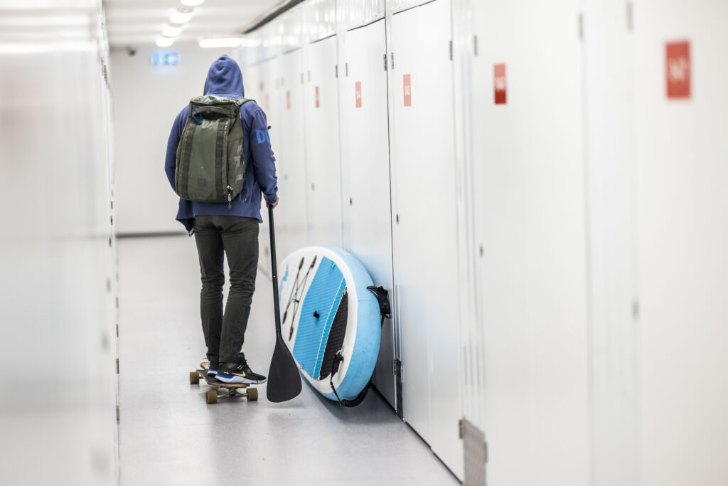 A boy is skating down a Flexistore facility with a SUP board next to him and a paddle in his hand. 